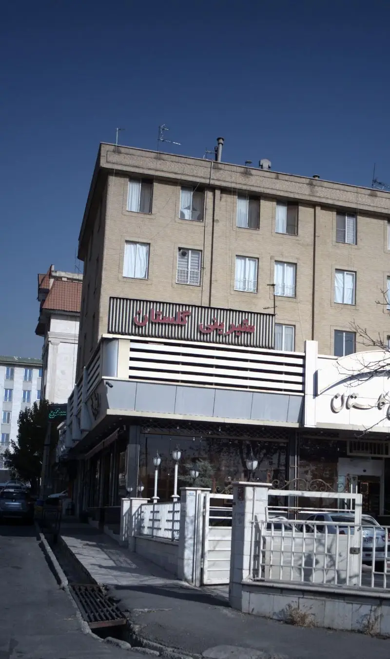 نمای بیرونی قنادی و شیرینی فروشی گلستان با تابلوی قرمز رنگ، در طبقه همکف ساختمانی آجری با پنجره‌های متعدد در یک روز روشن و آفتابی.