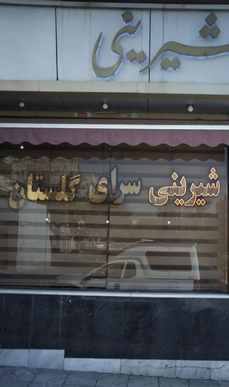 تصویر نمای بیرونی مغازه شیرینی گلستان با تابلوهای طلایی روشن، یک قنادی شیک که انواع کیک و شیرینی تازه را عرضه می‌کند.