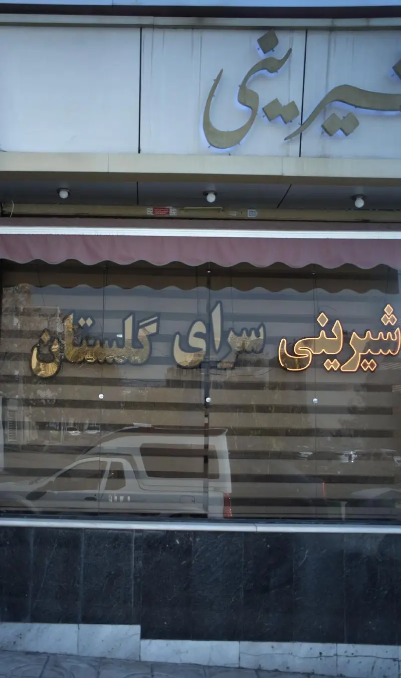 نمای خارجی شیرینی سرای گلستان با تابلوهای درخشان و حروف طلایی. بازتاب ماشین‌ها و خیابان در شیشه‌های مغازه دیده می‌شود.