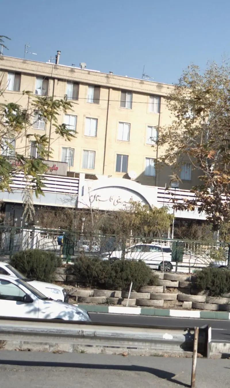 نمای خیابانی و ورودی شیرینی گلستان، قنادی معروف با تابلوی قوسی سفید رنگ در یک روز آفتابی، با خودروها و درختان.