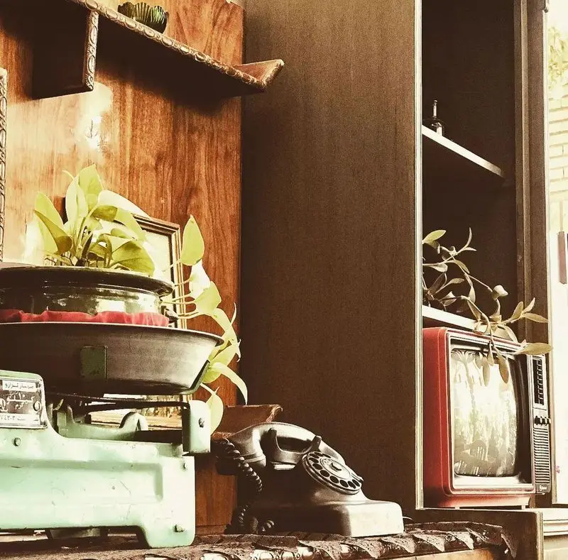 فضای داخلی دنج و نوستالژیک کافه دلواز با دکوری سنتی شامل ترازوی قدیمی، تلفن رومیزی وینتیج، تلویزیون قرمز و گلدان طبیعی.