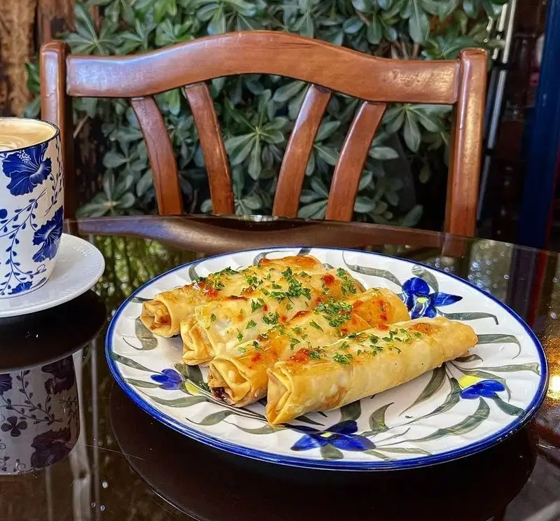 رول‌های پنیری طلایی و خوش‌طعم کافه دلواز، با سس مخصوص و جعفری روی بشقابی هنری، همراه با یک لاته آرت زیبا در فضایی دلنشین و سرسبز کافه