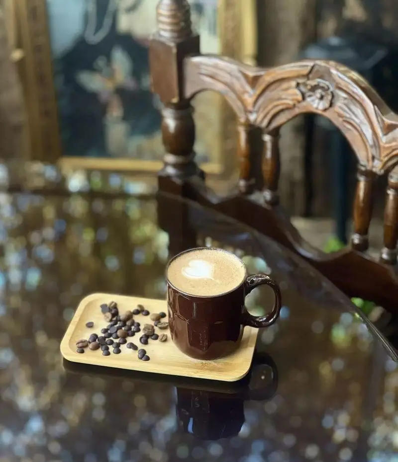 کافی لاته گرم با لاته آرت قلب، دانه قهوه و شکلات در سینی چوبی کافه دلواز؛ لحظاتی آرامش‌بخش بر روی میز شیشه‌ای.