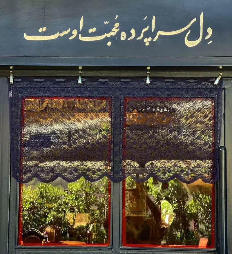 کافه دلواز: نمای جذاب ورودی با تابلو خوشنویسی طلایی شعر "دل سر اپرده محبت اوست" بالای پنجره‌های قرمز و پرده‌های توری ظریف.