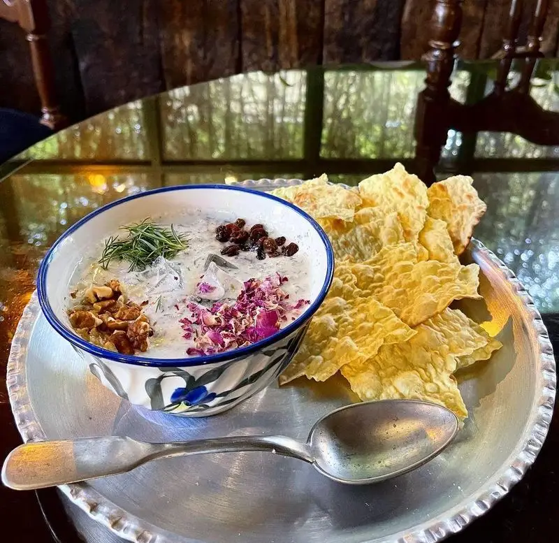 کاسه آبدوغ خیار خنک و سنتی کافه دلواز با گردو، گل محمدی، سبزی تازه و نان خشک محلی در سینی نقره‌ای