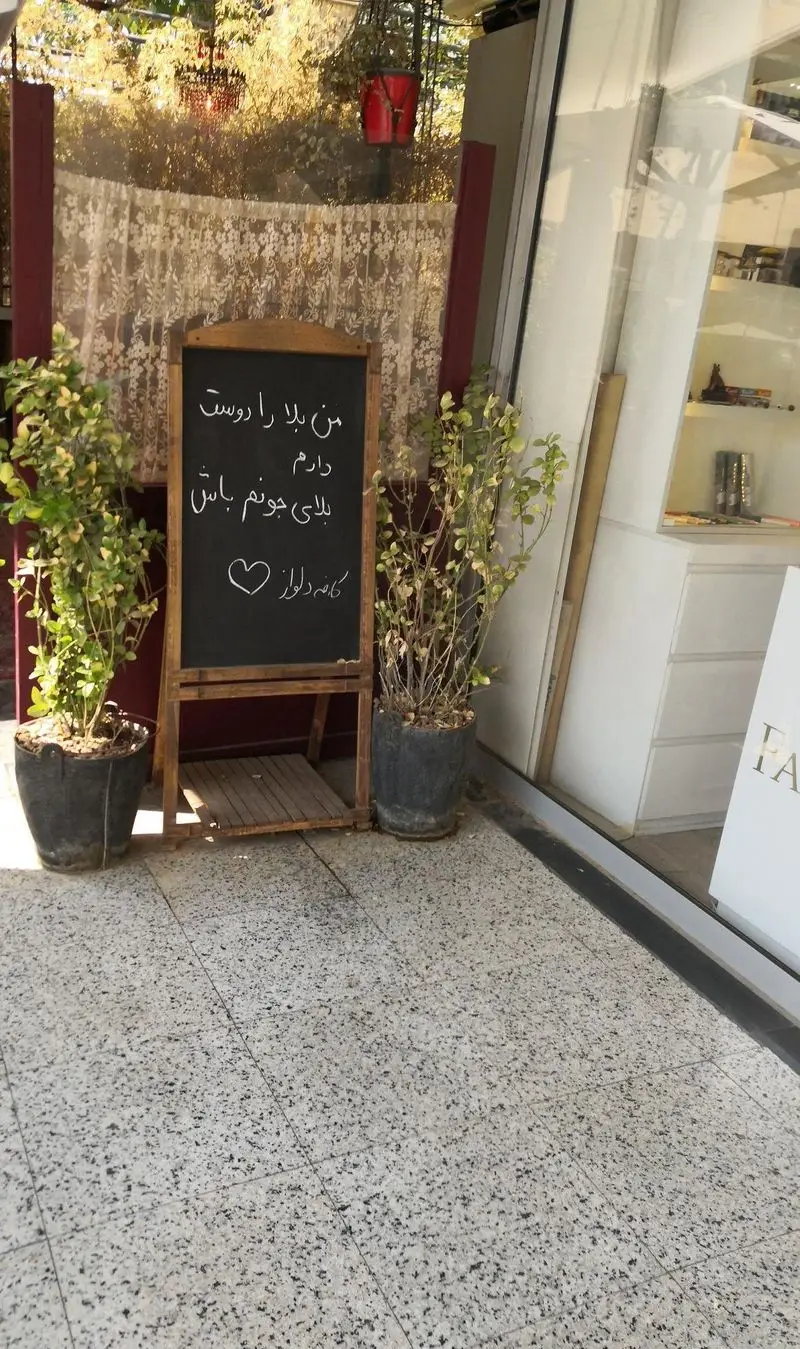 ورودی دلنشین کافه دلواز با تابلو گچی حاوی پیام عاشقانه و گلدان‌های سبز، فضای دنج و دعوت‌کننده‌ای را نشان می‌دهد.