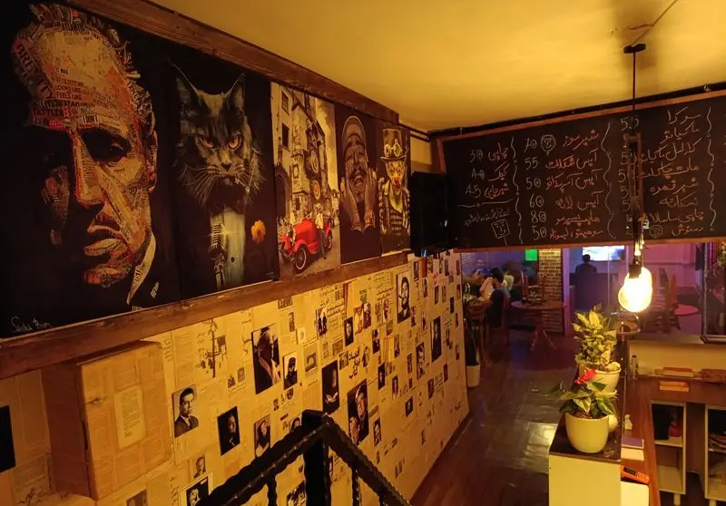 فضای داخلی کافه گیم شو، کافه کتاب مافیا گیم نت با دکوراسیون خاص، پوستر پدرخوانده، گربه مافیا و منوی متنوع روی تخته سیاه.