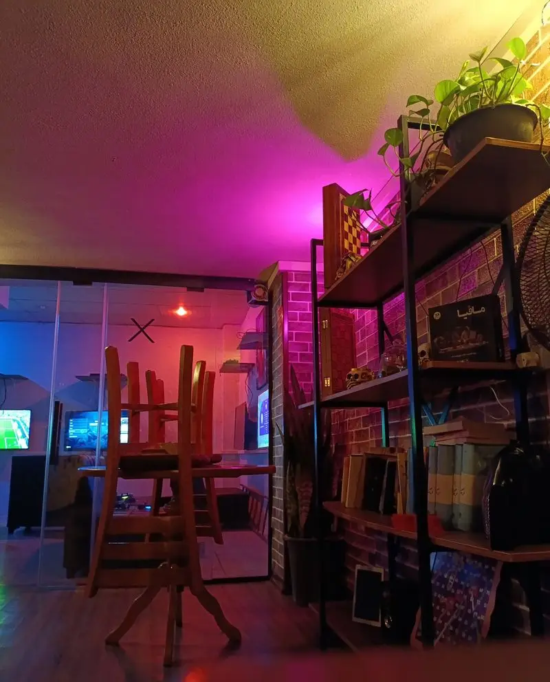 فضای داخلی کافه گیم و کتاب مافیا، با میز و صندلی چوبی، قفسه کتاب و بازی، نورپردازی خاص و دنج برای گیمرها و مطالعه.