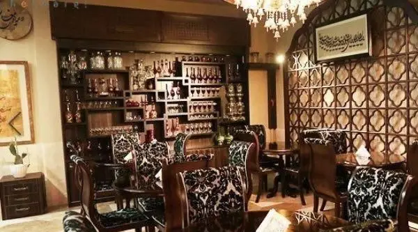 فضای داخلی شیک و سنتی رستوران ایرانی هزاردستان با دکوراسیون چوبی، قفسه‌های پر از نوشیدنی، میز و صندلی‌های طرح‌دار، و هنر خطاطی.