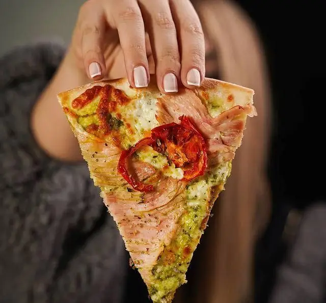 تصویری از یک تکه پیتزای لذیذ در دست، از رستوران ایتالیایی رومانا. پیتزای خاص با سس پستو، ژامبون و گوجه‌فرنگی خشک. تجربه‌ای بی‌نظیر.