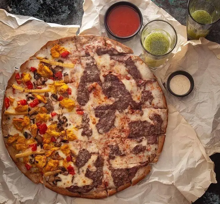 پیتزای دو طعم (نصف گوشت و نصف مرغ و سبزیجات شامل قارچ، ذرت و فلفل) از فست فود باگت با سس و نوشیدنی. مناسب برای لذت بردن از غذای خوشمزه.