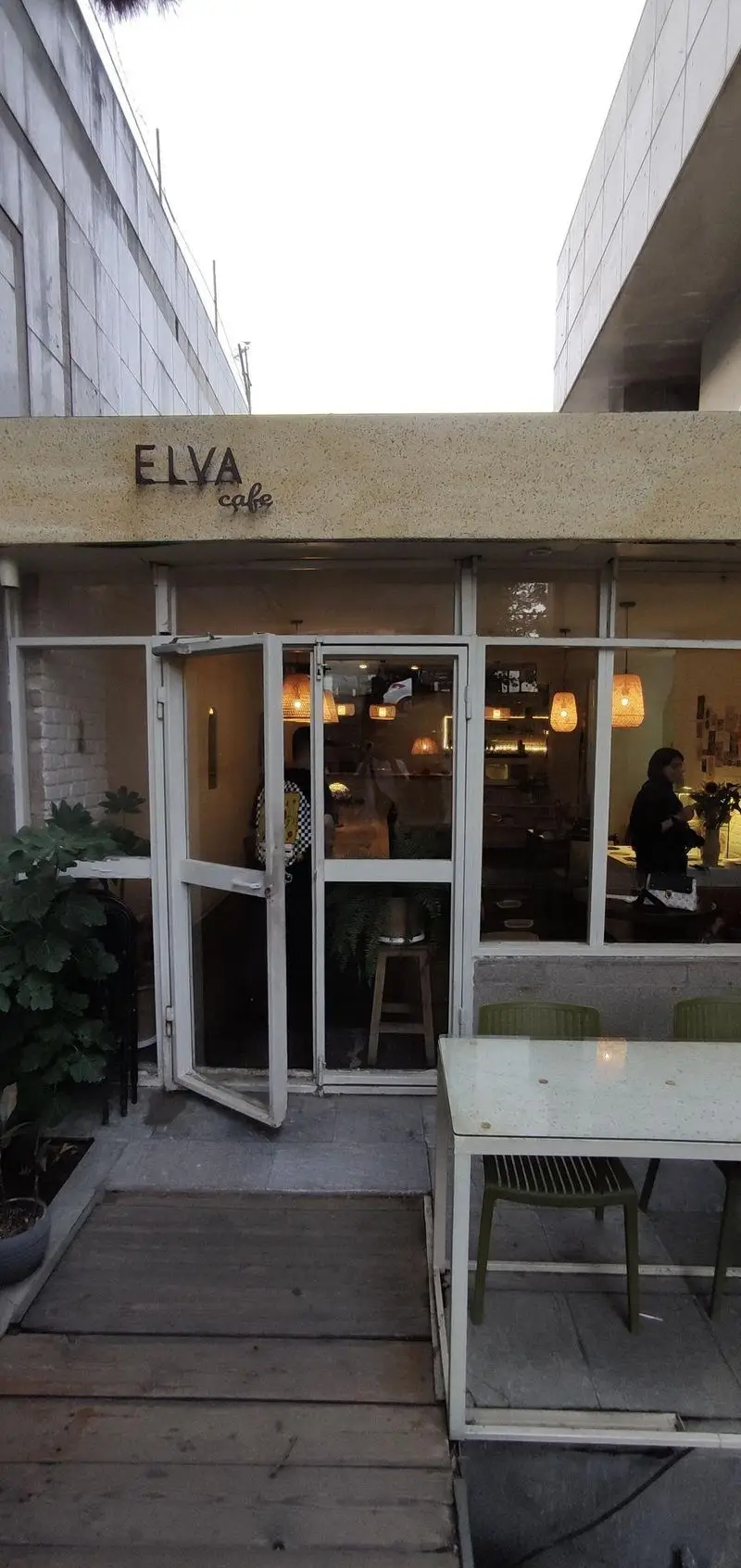 تصویری از نمای بیرونی کافه الوا با درب‌های شیشه‌ای بزرگ، فضای داخلی روشن با نورپردازی دکوراتیو و بخش نشیمن بیرونی با میز و صندلی.