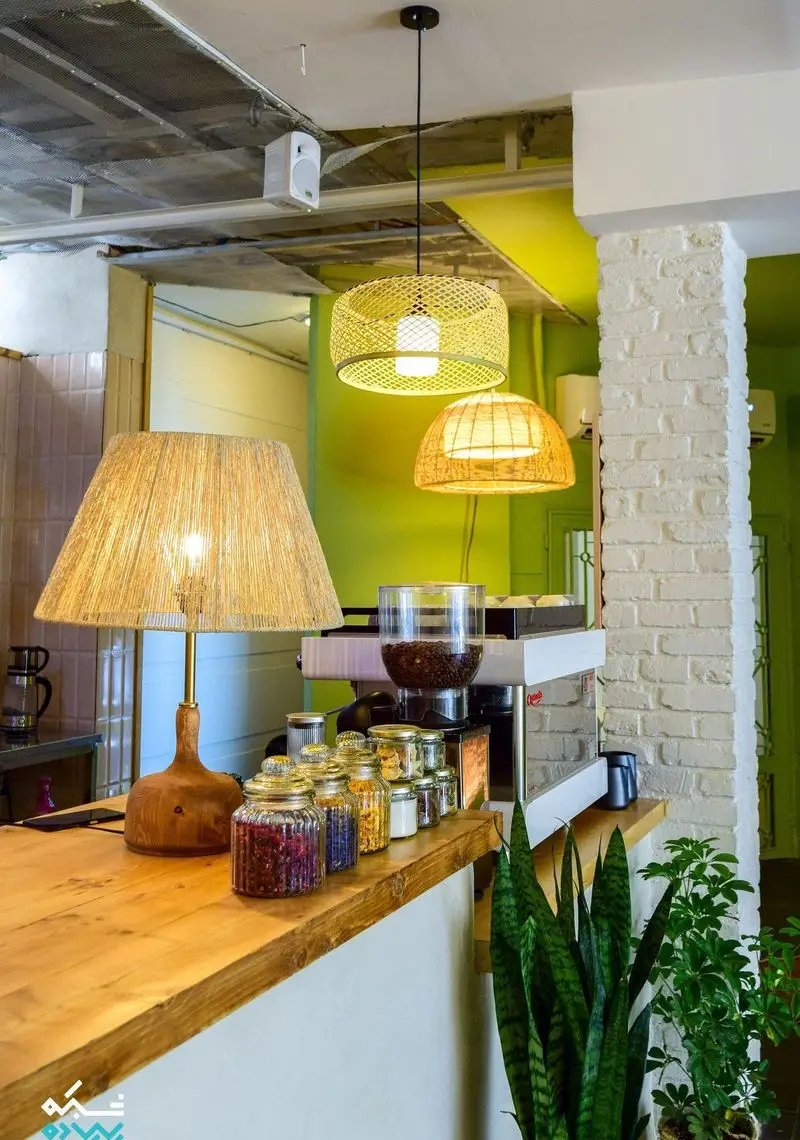 کافه الوا، دکوراسیون داخلی مدرن با میز چوبی بار، چراغ‌های آویز حصیری، قهوه‌ساز و ظروف شیشه‌ای رنگارنگ. فضایی گرم و دلنشین.