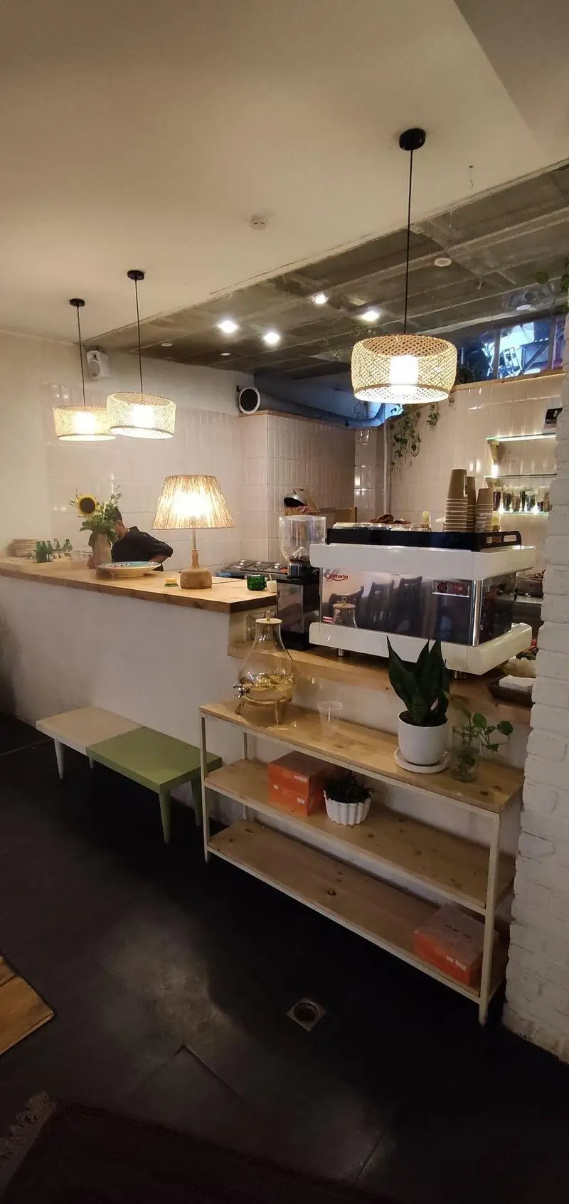 فضای داخلی کافه الوا؛ دکوراسیون مدرن و دنج با میز پیشخوان چوبی، دستگاه قهوه، نورپردازی گرم و چراغ‌های حصیری. محیطی آرام و دلنشین برای صرف قهوه.