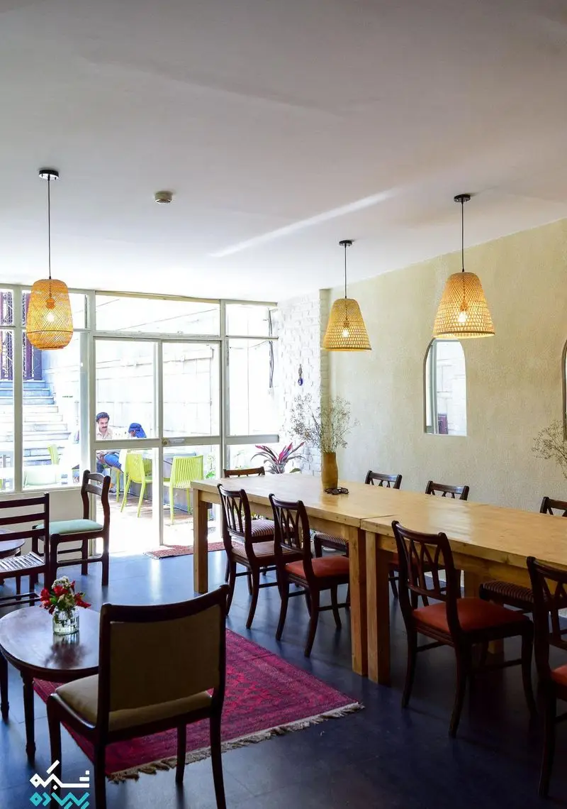 دکوراسیون داخلی دلنشین کافه الوا با میزهای چوبی بلند، صندلی‌های راحت، نورپردازی حصیری و چشم‌انداز فضای بیرونی. مکانی دنج برای دورهمی.