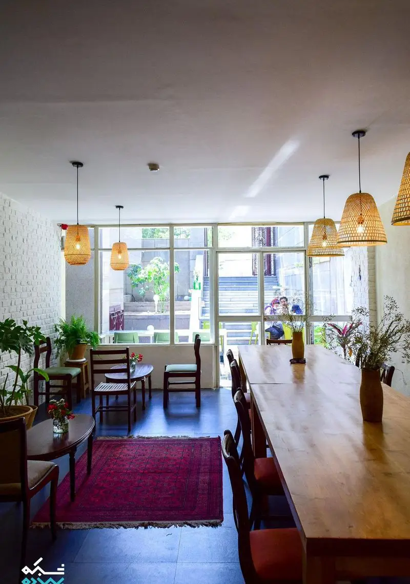 فضای داخلی کافه الوا با دکوراسیونی گرم و دنج: میزهای چوبی متنوع، صندلی‌های راحت، قالیچه ایرانی، گیاهان، نورپردازی حصیری و پنجره بزرگ.