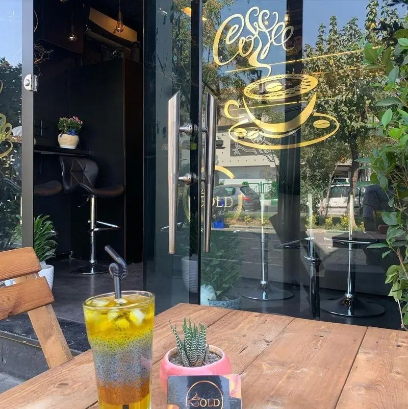 تصویری از کافه گلد: ورودی مدرن با لوگوی طلایی قهوه روی شیشه، دکوراسیون شیک و میز چوبی در فضای باز با نوشیدنی خنک.