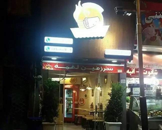 نمای شبانه و ورودی فست فود سرزمین لقمه با تابلوهای روشن و لوگوی ساندویچ. مکانی ایده آل برای سفارش لقمه‌ها و غذاهای خوشمزه.