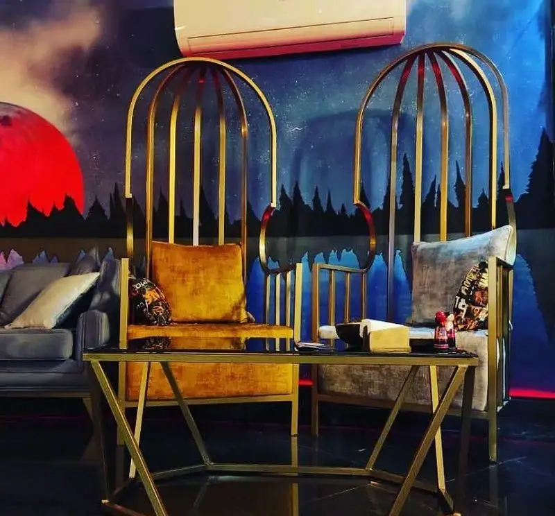 کافه شیرز: صندلی‌های طلایی شیک و دکوراسیون هنری دیوار با طرح ماه و جنگل، ایجاد فضایی دلپذیر برای مشتریان کافه.