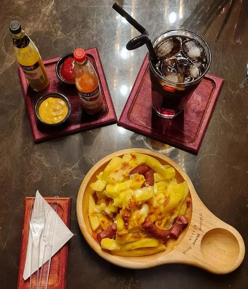 میز چوبی کافه شیرز با غذای خوشمزه: سیب زمینی پنیری و سوسیس، همراه با نوشیدنی خنک و انواع سس. فضایی دلنشین برای لذت بردن.