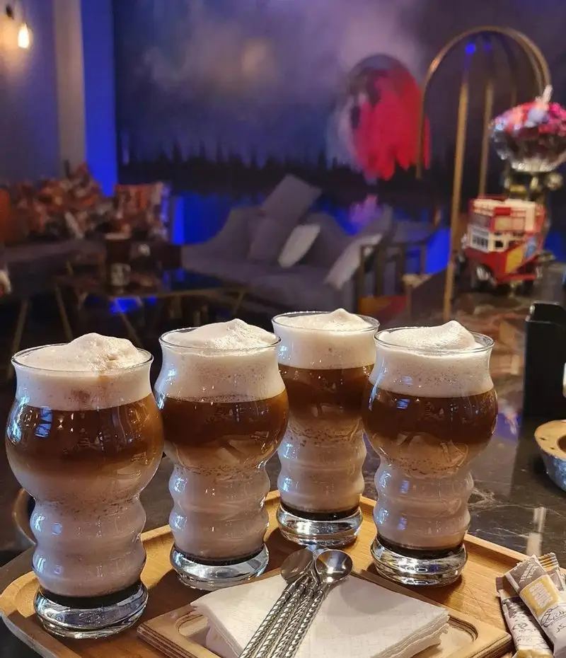 تصویری زیبا از چهار نوشیدنی لایه ای قهوه سرد یا لاته با فوم فراوان در لیوان های شیک، سرو شده در سینی چوبی کافه شیرز با دکوراسیون مدرن و نورپردازی آبی.