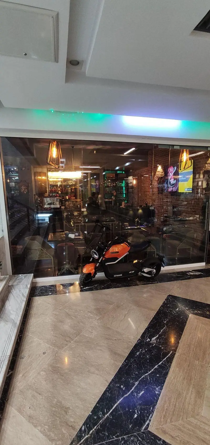 فضای ورودی کافه شیرز با پنجره‌های شیشه‌ای بزرگ، دکوراسیون داخلی مدرن و لوکس، نورپردازی جذاب و موتورسیکلت نارنجی جلوی آن.