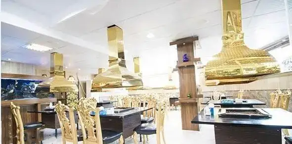 فضای داخلی رستوران اجاق باشی با میز و صندلی‌های طلایی لوکس و هودهای کباب‌پز طلایی بالای هر میز، به همراه آکواریوم بزرگ زیبا.