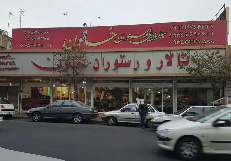 نمای بیرونی رستوران و تالار پذیرایی خاتون، تابلوی قرمز رنگ با نوشته‌های طلایی و شماره تلفن‌ها، در یک روز ابری.