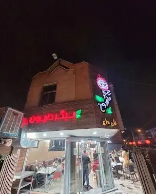نمای بیرونی کبابی جیگر طهرون در شب با نورپردازی و مشتریان. تابلوی نئون روشن "جیگر طهرون" و "نان داغ" به وضوح دیده می‌شود.