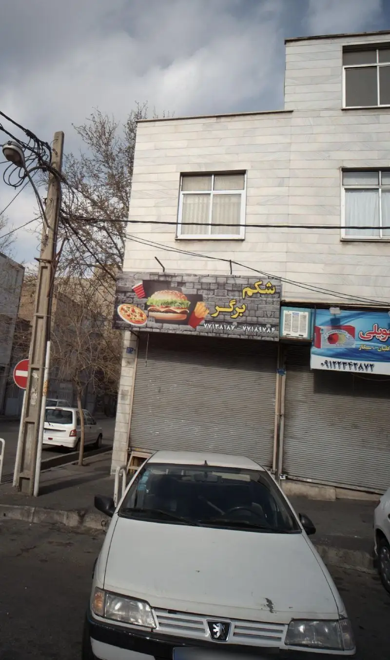 نمای بیرونی فست فود شکم برگر، تابلویی با تصویر برگر، پیتزا، سیب‌زمینی و نوشابه در خیابان. خودرویی سفید جلوی مغازه پارک شده.