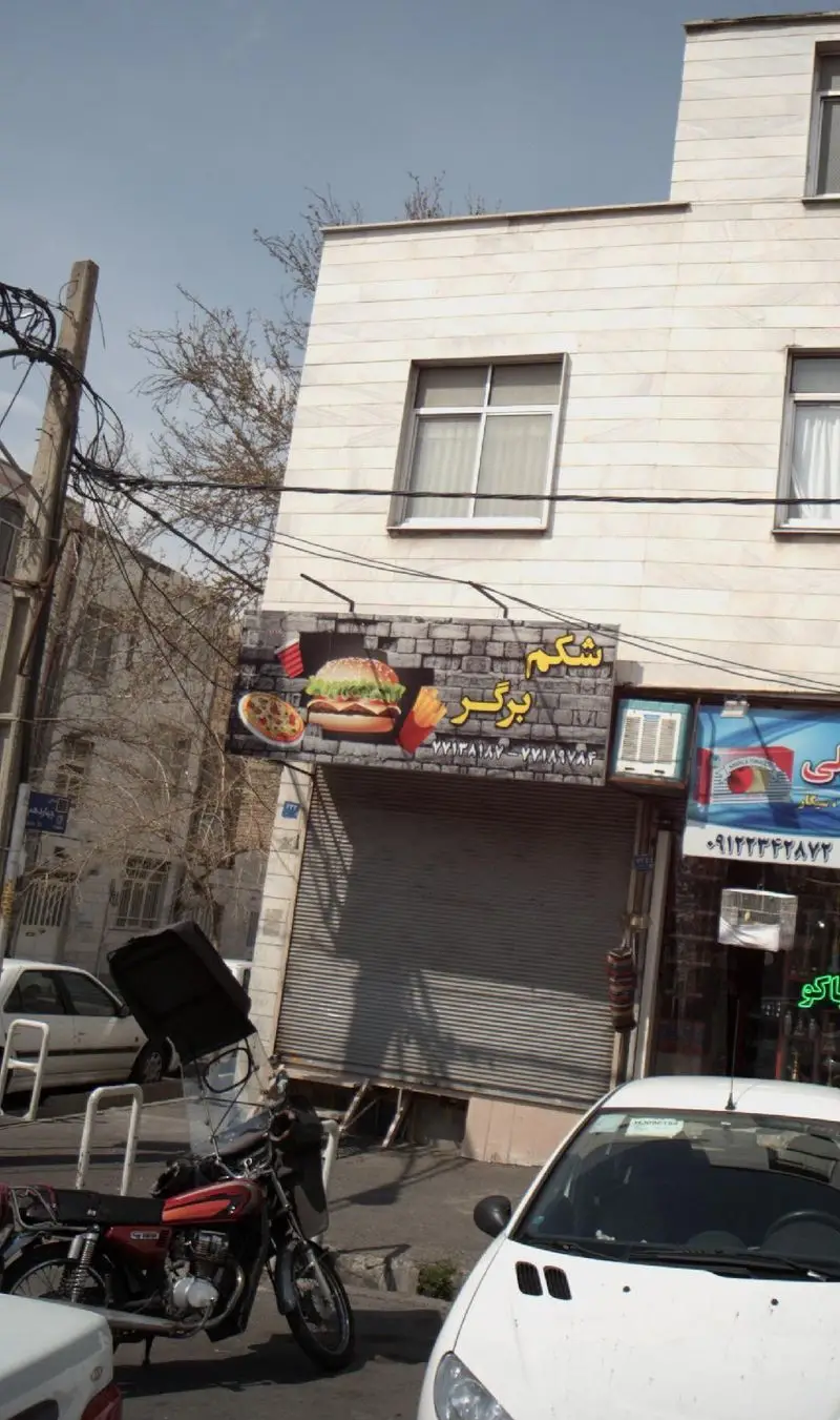 تصویری از نمای بیرونی فست فود شکم برگر، شامل تابلو با عکس‌های جذاب برگر، پیتزا، سیب زمینی و نوشابه در شهر تهران.