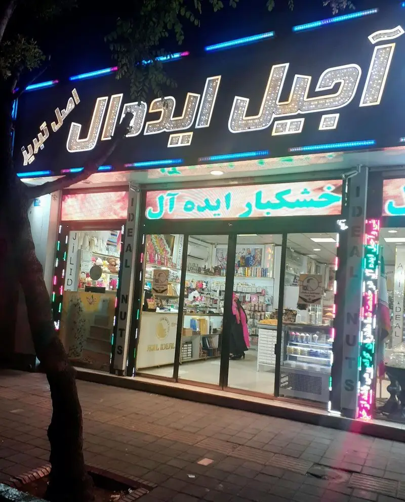 تصویر نمای بیرونی شبانه شیرینی و آجیل سرای ایده آل تبریز با تابلوهای درخشان و نورپردازی جذاب.