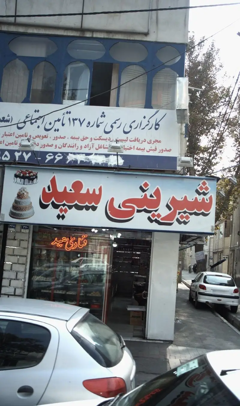 تصویری از نمای بیرونی شیرینی سعید، یک قنادی مجهز با انواع کیک و شیرینی‌های تازه و خوشمزه. دفتر تامین اجتماعی نیز در طبقه بالا پیداست.