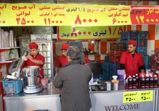 نمایی از ایستگاه آب‌ هویج و بستنی آرتین با مشتری و فروشندگان در حال آماده‌سازی آب‌میوه و بستنی سنتی، همراه با لیست قیمت آب هویج و آب انار.