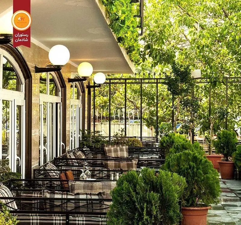فضای باز و دنج رستوران شادمان با میز و صندلی‌های چیدمان شده، پتوهای چهارخانه، گلدان‌های زیبا و درختان سرسبز، مکانی دلنشین برای مشتریان.