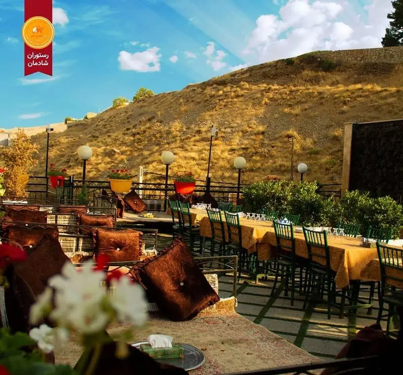رستوران شادمان با فضای باز دلنشین، میزهای چیده شده آماده پذیرایی از مهمانان، کوسن‌های راحت و منظره زیبای کوه در روز آفتابی