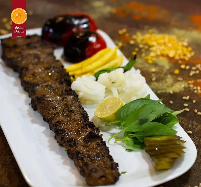 تصویری جذاب از کباب کوبیده سنتی رستوران شادمان به همراه گوجه کبابی، ترشی، سبزی تازه و لیمو. تجربه غذای اصیل ایرانی.