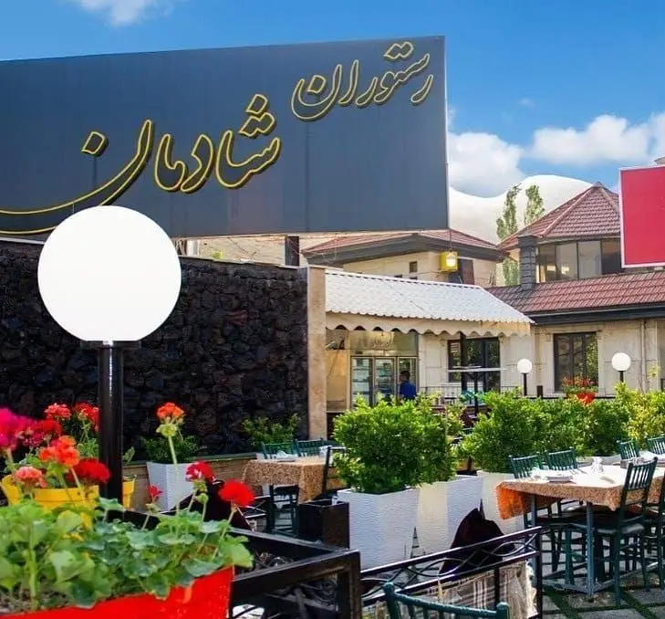 نمایی زیبا از رستوران شادمان با تابلو طلایی، فضای باز سرسبز و دلنشین، میز و صندلی‌های چیده شده آماده پذیرایی از میهمانان.