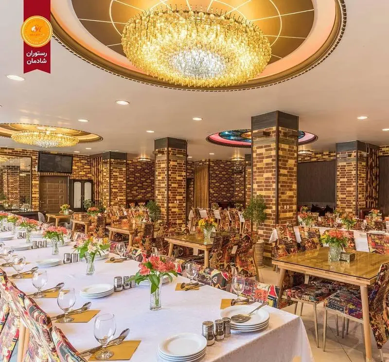 فضای داخلی مجلل و دلباز رستوران شادمان، با میزهای آماده پذیرایی، صندلی‌های طرح‌دار، دیوارهای آجری و لوسترهای کریستالی در سقف.