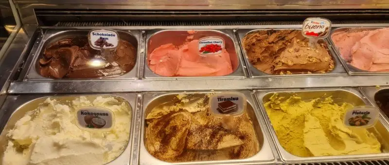 نمایش انواع بستنی‌های دست‌ساز و خوش‌طعم در بستنی سان مار کو شامل شکلات رام، گیلاس، کیندر بوئنو، نارگیل، تیرامیسو، و پسته.