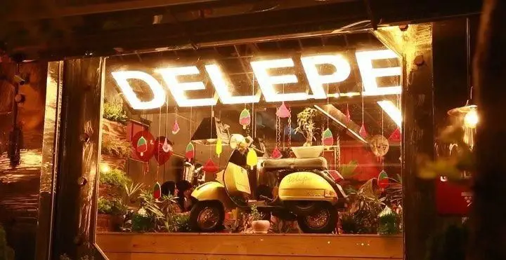 تصویر شب از نمای بیرونی فست فود و کافه د ل پ، با تابلوی روشن "DELEP" و اسکوتر زرد رنگ و دکوراسیون داخلی جذاب در ویترین آن.