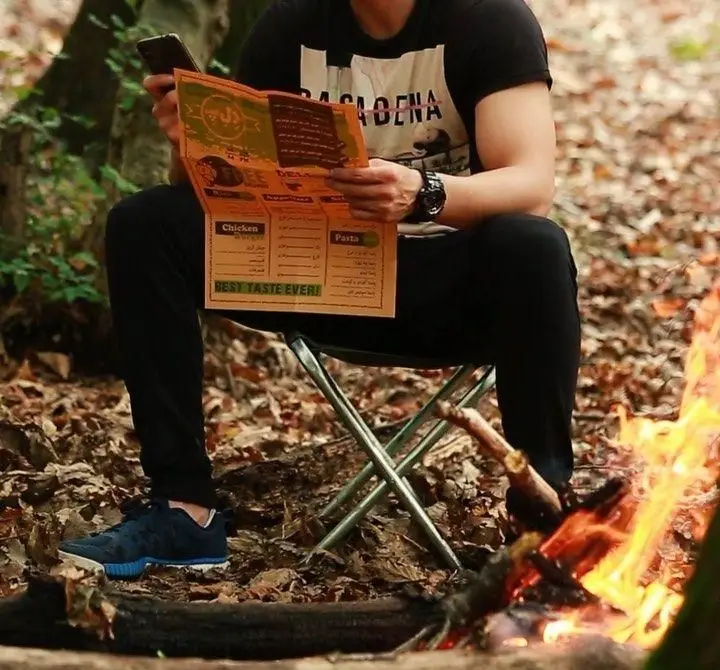 مردی در طبیعت کنار آتش، منوی فست فود د ل پ را برای انتخاب غذای خوشمزه مشاهده می‌کند. سفارش غذای بیرون بر با بهترین طعم برای کمپینگ.
