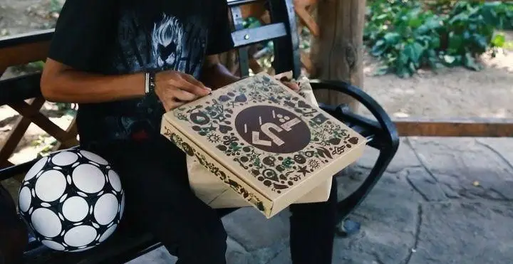 تجربه لذت‌بخش سفارش فست فود د ل پ: جعبه پیتزایی با طراحی جذاب و لوگوی منحصربه‌فرد در محیطی آرام و دلپذیر، کنار توپ فوتبال.