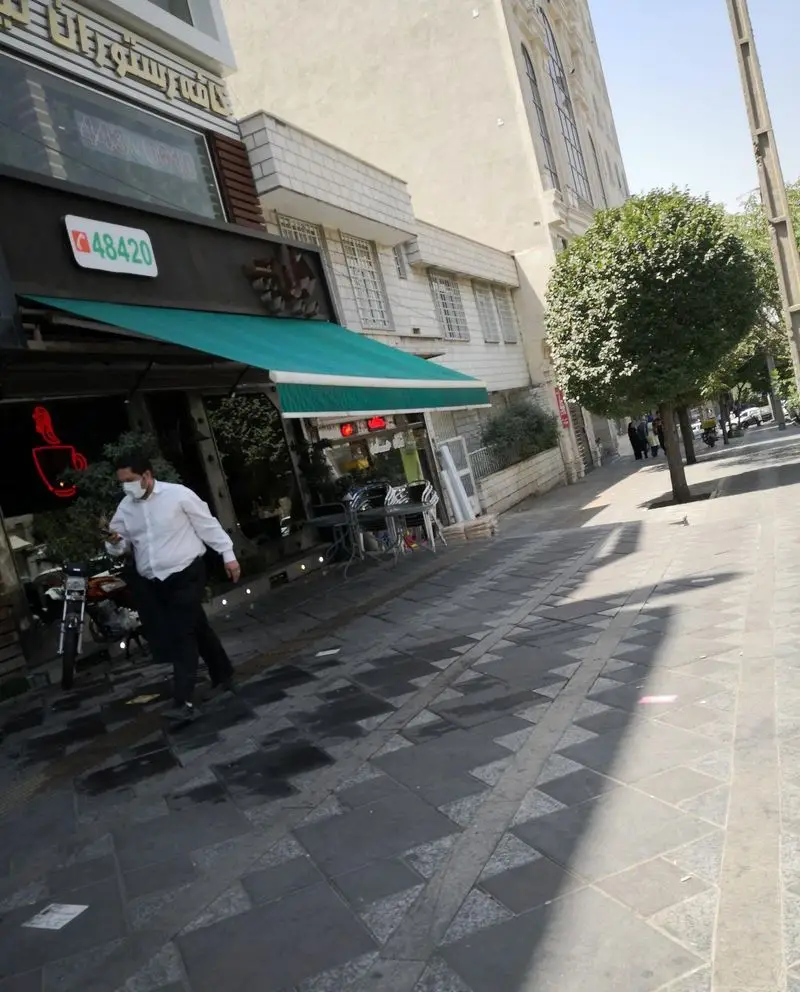 نمای خیابان از فست فود د ل پ با تابلوی سبز، فضای نشیمن بیرونی، پیاده‌رو طرح‌دار، و مردی با ماسک در حال عبور.