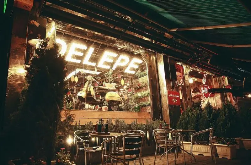 نمای خارجی شبانه فست فود د ل پ با تابلو نئون DELEPE روشن، دکوراسیون منحصر به فرد شامل اسکوتر و فضای باز دلنشین.