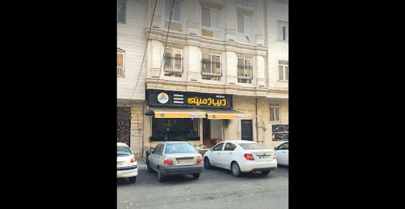 نمای خارجی شعبه دیب دمینی سعادت آباد، یک فست فود یا رستوران با تابلوی برند زرد رنگ روشن و چند خودرو در جلوی آن.
