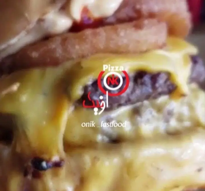 نمایی نزدیک و هوس‌انگیز از یک برگر آبدار با پنیر ذوب شده فراوان و حلقه‌های پیاز ترد در فست فود اونیک. لوگوی پیتزا NK نیز مشهود است.