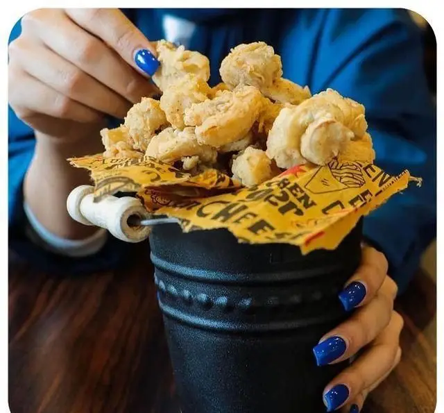 یک سطل کامل پاپ کورن چیکن طلایی و ترد از فست فود اونیک، با کاغذ زرد مارک دار. دستی با لاک آبی در حال برداشتن یک تکه خوشمزه.