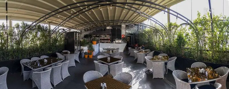 فضای باز رستوران بنلی لانژ با میز و صندلی حصیری مدرن، سقف متحرک، بار نوشیدنی و محیطی سبز و دلنشین برای صرف غذا و قهوه.