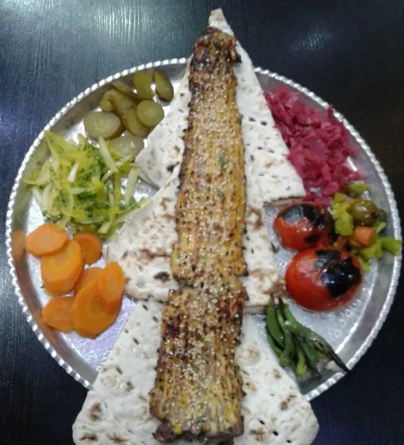 یک پرس کباب کوبیده یا لقمه مخصوص با کنجد، نان تازه، گوجه کبابی، انواع ترشیجات و سالاد در رستوران سنتی دنج.